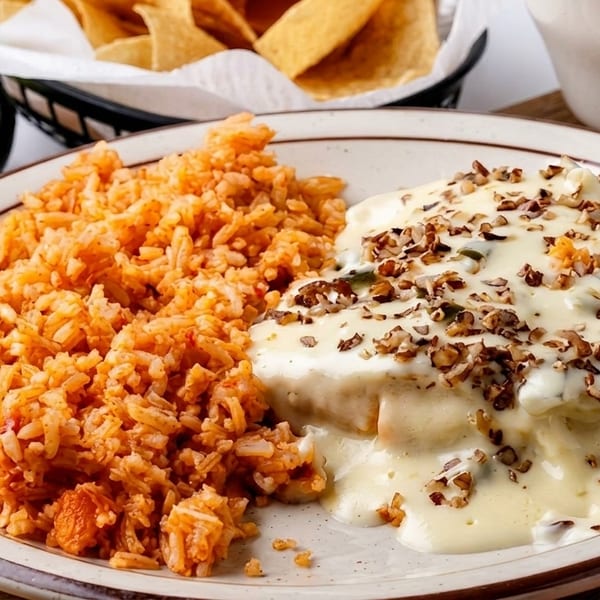 A plate of Mexican food with red rice and a dish covered in white sauce and nuts.